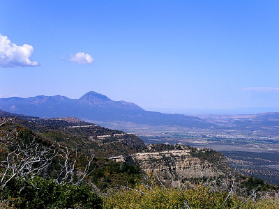 [Vista view with two levels of mountains. In the foreground coming from the left is a rocky incline which is covered in greenery at the top. In the middle ground on the right is the valley floor. In the far distance is a row of mountains with a big hump (Sleeping Ute)in the middle.]